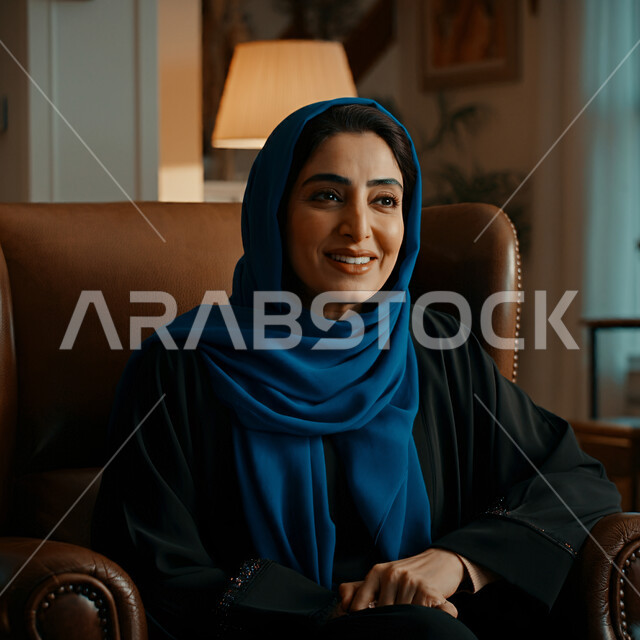 قضاء اوقات ممتعة في المنزل، الهدوء والاسترخاء والراحة، أزياء محجبات عصرية راقية، مفهوم الأناقة والاهتمام بالمظهر الخارجي، صورة مقربة لامرأة عربية خليجية سعودية مبتسمة ترتدي العباءة والحجاب تنظر الى شيء ما بإيماءات السرور و الثقة بالنفس