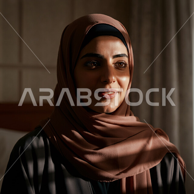 أزياء محجبات عصرية راقية، الاهتمام بالمظهر الخارجي، قضاء اوقات ممتعة في المنزل، مفهوم الهدوء والاسترخاء والراحة، صورة مقربة لامرأة عربية خليجية سعودية مبتسمة ترتدي العباءة والحجاب تنظر الى شيء ما بإيماءات السرور و الثقة بالنفس