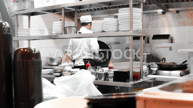 احتراف مهنة وفنون الطبخ، صورة من الخلف لشيف عربي خليجي سعودي يرتدي الكمامة وزي الطهاة يقوم باعداد وتجهيز وجبة الطعام على موقد غاز، الفن في اعداد وتقديم الاصناف المتنوعة من الاطعمة والمأكولات الشرقية اللذيذة، اتباع اجراءات السلامة في مطابخ ومطاعم المملكة العربية السعودية