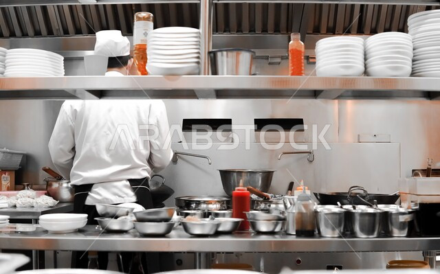 احتراف مهنة وفنون الطبخ، صورة من الخلف لشيف عربي خليجي سعودي يرتدي الكمامة وزي الطهاة يقوم باعداد وتجهيز وجبة الطعام على موقد غاز، الفن في اعداد وتقديم الاصناف المتنوعة من الاطعمة والمأكولات الشرقية اللذيذة، اتباع اجراءات السلامة في مطابخ ومطاعم المملكة العربية السعودية