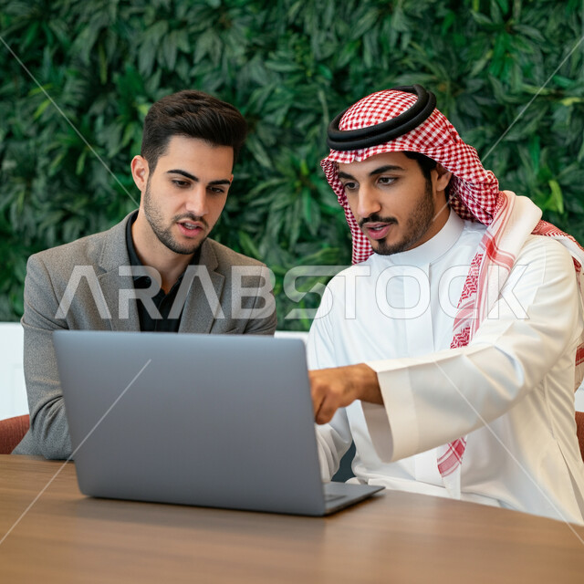 مفهوم الشراكة والتعاون وتبادل الخبرات والمهارات، موظفان عربيان خليجيان سعوديان يعملان داخل المكتب، استخدام الأجهزة والتقنيات الحديثة في الأعمال المكتبية، إنجاز وتحقيق مهام العمل، شاب يشير بإصبع السبابة إلى شاشة الحاسوب