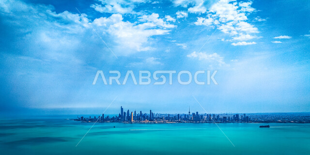 الواجهة البحرية لمدينة الكويت، صورة بطائرة دون طيار للأبراج وناطحات السحاب والمنشآت والمباني التجارية المطلة على ساحل الخليج العربي، مياه البحر الزرقاء الصافية في وقت النهار مع الغيوم البيضاء في السماء، خلفية الطبيعة