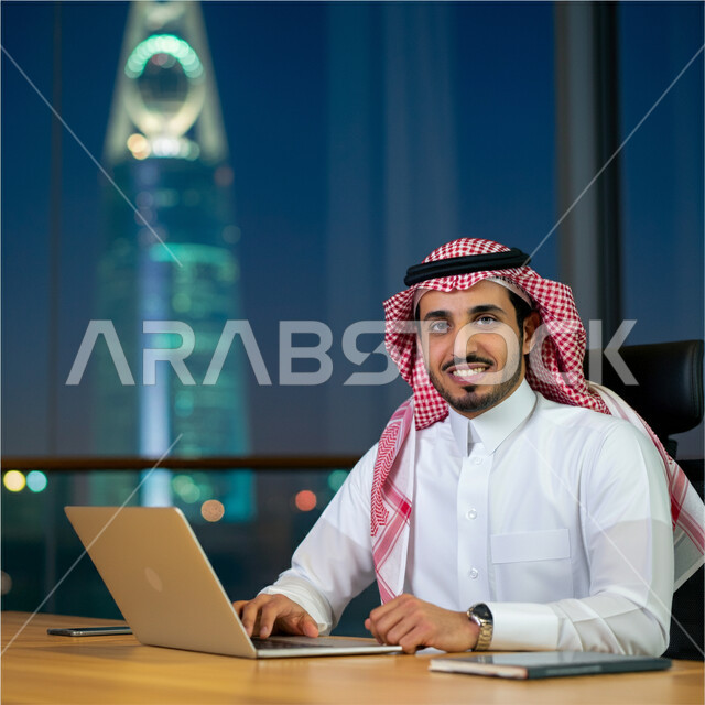 دمج العمل مع التقنية والتكنولوجيا، إدارة شؤون الأعمال وتنظيمها عبر الأجهزة التقنية، صورة مقربة لرجل عربي خليجي سعودي يرتدي الشماغ و الثوب التقليدي يعمل على الحاسوب المحمول داخل مقر الشركة، مهن ووظائف إدارية مكتبية في المملكة العربية السعودية