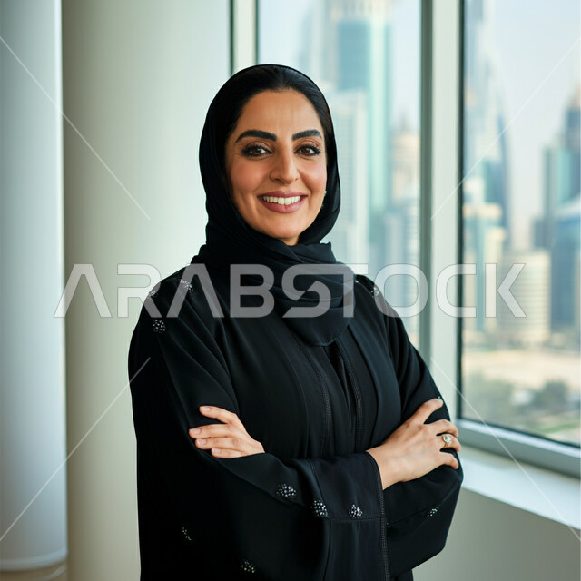أزياء محجبات عصرية راقية، الأناقة والاهتمام بالمظهر الخارجي، صورة مقربة لامرأة عربية خليجية سعودية مبتسمة ترتدي الحجاب والعباءة السوداء تقف بأيدي متقاطعة تنظر إلى الكاميرا بإيماءات السرور والثقة بالنفس، مفهوم النعومة والأنوثة