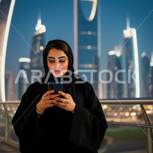 استخدام الأجهزة التقنية الحديثة، مفهوم النعومة والأنوثة، صورة مقربة لامرأة عربية خليجية سعودية ترتدي الحجاب والعباءة السوداء تمسك بيدها الهاتف المحمول، التقاط الصور التذكارية وسط المدينة، التواصل مع الأهل والأصدقاء، خلفية الأبراج وناطحات السحاب