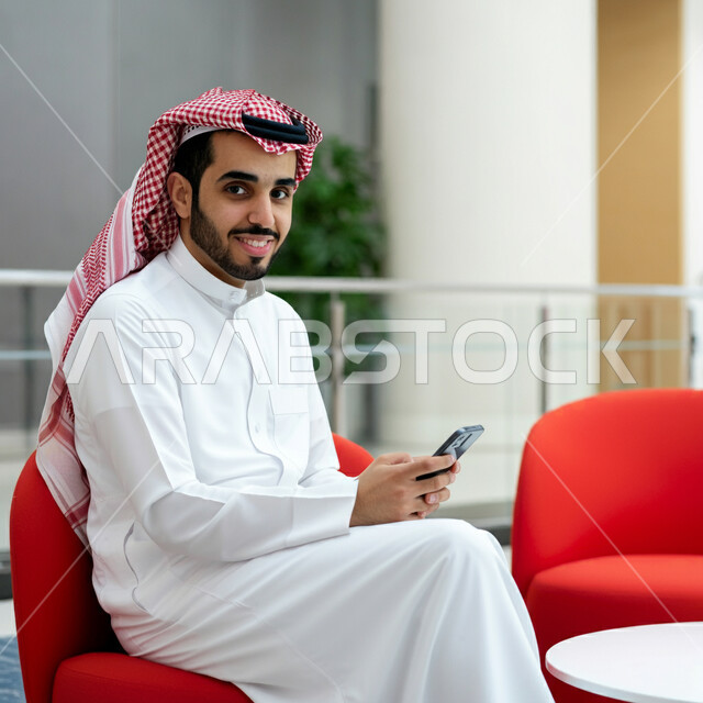 التواصل والدردشة مع الأهل والأصدقاء، رجل عربي خليجي سعودي يرتدي الثوب التقليدي والشماغ يجلس على كرسي مريح يمسك بيده الهاتف المحمول ينظر إلى الكاميرا بإيماءات السرور، استخدام الأجهزة التقنية الحديثة، إجراء مكالمات الفيديو عبر الموبايل