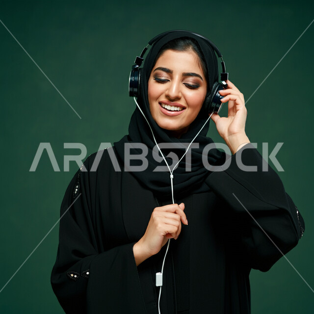استخدام الهيدفون في اوقات الفراغ، بورتريه مقرب لإمرأه عربيه سعوديه خليجيه محجبة ترتدي عباءة سوداء مع سماعة الرأس بايماءات الإندماج والتفاعل وسماع الموسيقى، مفهوم التسليه والترفيه, قضاء وقت ممتع, تطور التقنيه والتكنولوجيا، خلفية خضراء