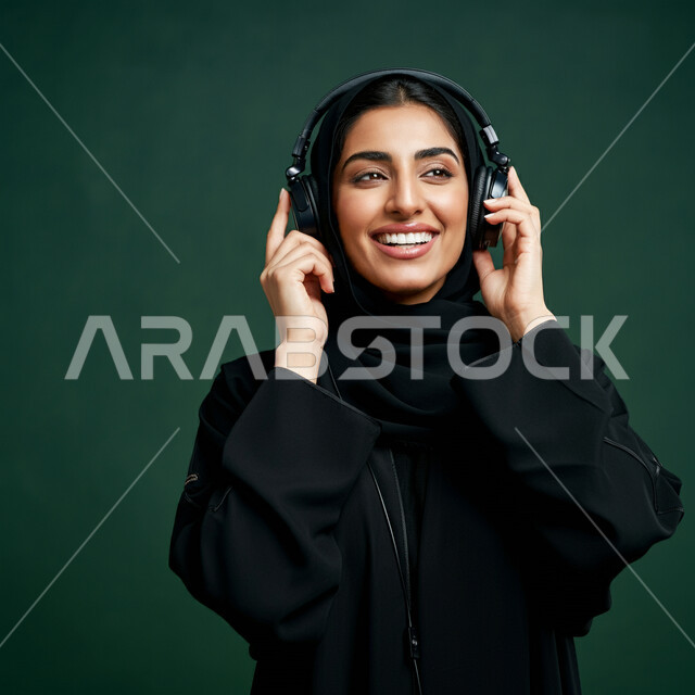 Using headphones in free time, close-up portrait of a veiled Saudi Arabian Gulf woman wearing a black abaya with headphones with gestures of integration, interaction and listening to music, the concept of entertainment and leisure, having a good time, development of technology and technology, green background