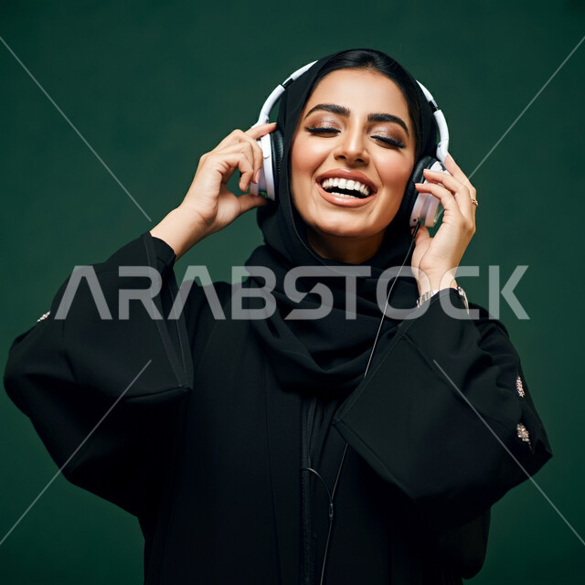 قضاء وقت ممتع, استخدام الهيدفون في اوقات الفراغ، بورتريه مقرب لإمرأه عربيه سعوديه خليجيه محجبة ترتدي عباءة سوداء مع سماعة الرأس بايماءات الإندماج والتفاعل وسماع الموسيقى، مفهوم التسليه والترفيه, تطور التقنيه والتكنولوجيا، خلفية خضراء