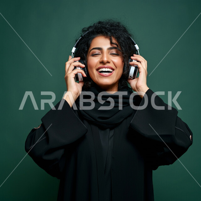 قضاء وقت ممتع, استخدام الهيدفون في اوقات الفراغ، بورتريه مقرب لإمرأه عربيه سعوديه خليجيه محجبة ترتدي عباءة سوداء مع سماعة الرأس بايماءات الإندماج والتفاعل وسماع الموسيقى، مفهوم التسليه والترفيه, تطور التقنيه والتكنولوجيا، خلفية خضراء