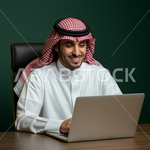 استخدام الأجهزة التقنية الحديثة، إدارة شؤون الأعمال وتنظيمها، بورتريه مقرب لرجل عربي خليجي سعودي يرتدي الشماغ و الثوب التقليدي يجلس على كرسي مريح يعمل على الحاسوب المحمول، مهن ووظائف إدارية، مفهوم إنجاز المشاريع والمهام، العمل عن بعد، خلفية خضراء