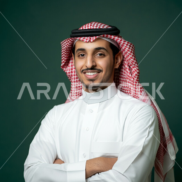 The concept of elegance and masculinity, self-confidence and interest in appearance, close-up portrait of a young Saudi Arabian Gulf man wearing traditional thobe and shemagh looking at the camera with expressions of happiness and pleasure, standing with crossed hand gestures, green background