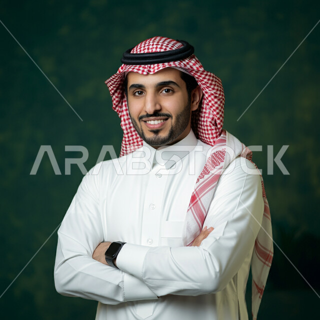 The concept of elegance and masculinity, self-confidence and interest in appearance, close-up portrait of a young Saudi Arabian Gulf man wearing traditional thobe and shemagh looking at the camera with expressions of happiness and pleasure, standing with crossed hand gestures, green background