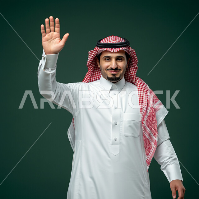 النظر الى الكاميرا بايماءات السعادة والسرور، مفهوم الاستقبال والترحيب، التعارف و الصداقه وتكوين علاقات, إلقاء التحيه والسلام, بورتريه مقرب لرجل عربي خليجي سعودي يرتدي الثوب التقليدي والشماغ يقوم برفع كفه,مفهوم الاناقه و الرجولة، الاهتمام بالمظهر الخارجي، خلفية خضراء