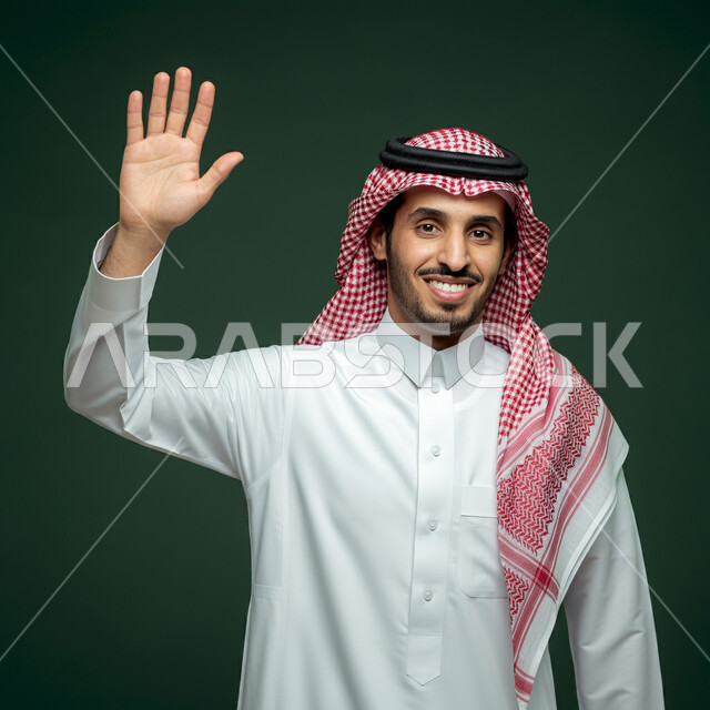 النظر الى الكاميرا بايماءات السعادة والسرور، مفهوم الاستقبال والترحيب، التعارف و الصداقه وتكوين علاقات, إلقاء التحيه والسلام, بورتريه مقرب لرجل عربي خليجي سعودي يرتدي الثوب التقليدي والشماغ يقوم برفع كفه,مفهوم الاناقه و الرجولة، الاهتمام بالمظهر الخارجي، خلفية خضراء