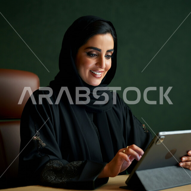Using modern technical devices, the concept of completing projects and tasks, working remotely, browsing social networking sites, communicating with family and friends via iPad, close-up portrait of a veiled Saudi Arabian Gulf woman wearing a black abaya using a tablet, green background