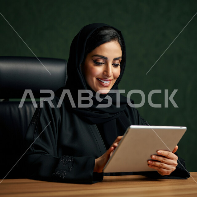 Using modern technical devices, the concept of completing projects and tasks, working remotely, browsing social networking sites, communicating with family and friends via iPad, close-up portrait of a veiled Saudi Arabian Gulf woman wearing a black abaya using a tablet, green background