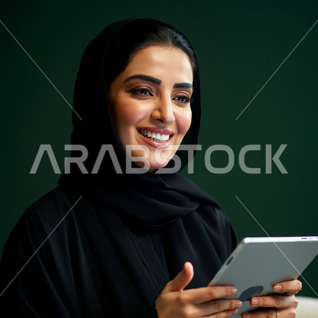 Using modern technical devices, the concept of completing projects and tasks, working remotely, browsing social networking sites, communicating with family and friends via iPad, close-up portrait of a veiled Saudi Arabian Gulf woman wearing a black abaya using a tablet, green background