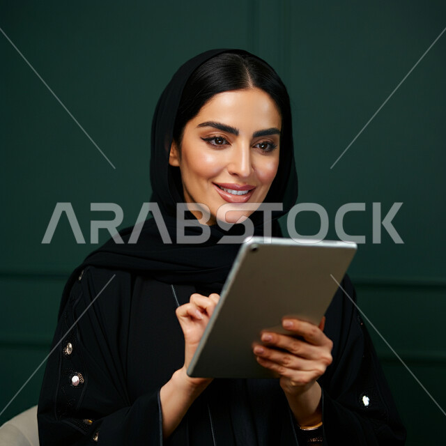 Using modern technical devices, the concept of completing projects and tasks, working remotely, browsing social networking sites, communicating with family and friends via iPad, close-up portrait of a veiled Saudi Arabian Gulf woman wearing a black abaya using a tablet, green background