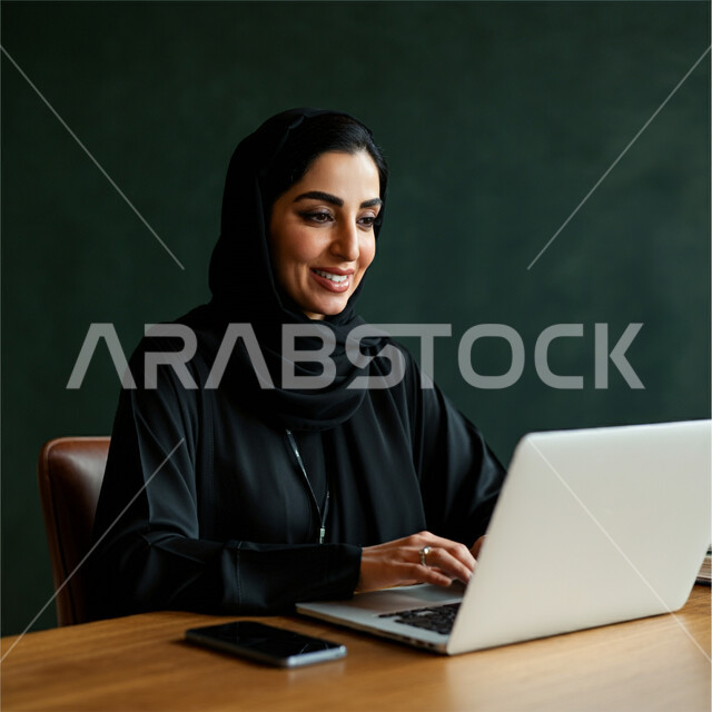 Concept of completing projects and tasks, using modern technical devices, managing and organizing business affairs, close-up portrait of a veiled Saudi Arabian Gulf woman wearing an abaya sitting on a comfortable chair working on a laptop, administrative professions and jobs, remote work, green background