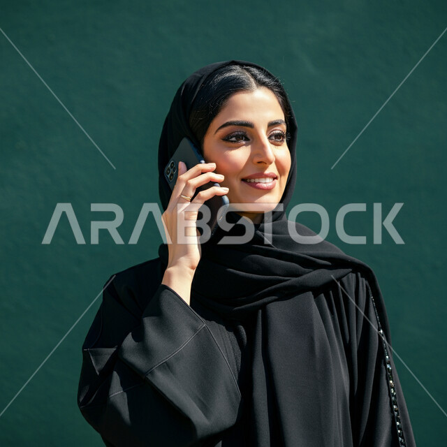 الإجابة والرد على الاتصالات بإستخدام الهاتف المحمول، بورتريه مقرب لامرأة عربية سعودية خليجية محجبة تقوم بإجراء مكالمة صوتية عبر الجوال، تعابير الفرح عند سماع الاخبار السارة، قضاء اوقات ممتعة بتبادل الاخبار والأحاديث، التواصل والاطمئنان على الأصدقاء، خلفية خضراء