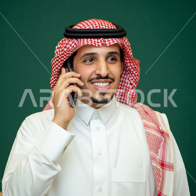 الإجابة والرد على الاتصالات بإستخدام الهاتف المحمول، بورتريه مقرب لرجل عربي سعودي خليجي يرتدي الثوب التقليدي يقوم بإجراء مكالمة صوتية، تعابير الفرح عند سماع الاخبار السارة، قضاء اوقات ممتعة بتبادل الاخبار والأحاديث، التواصل والاطمئنان على الأصدقاء، خلفية خضراء