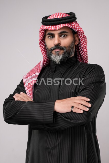 ايماءات الثقة بالنفس، مفهوم الاهتمام بالمظهر الخارجي و الرجولة والأناقه، بورتريه لرجل كبير بالسن عربي خليجي سعودي يرتدي الشماغ والزي التقليدي يقف باستقامة بايدي متقاطعه, النظر إلى الكاميرا بتعابيرالجدية والحزم، خلفية بيضاء