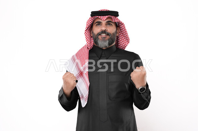 مفهوم القوة والانتصار وتحقيق الاهداف، ايماءات تدل على السعادة والحماس، بورتريه لرجل مسن عربي خليجي سعودي يرتدي الزي التقليدي والشماغ  يرفع يديه بكفين مقبوضتين, خلفية بيضاء