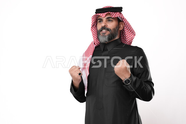 ايماءات تدل على السعادة والحماس، مفهوم القوة والانتصار وتحقيق الاهداف، بورتريه لرجل مسن عربي خليجي سعودي يرتدي الزي التقليدي والشماغ  يرفع يديه بكفين مقبوضتين, خلفية بيضاء