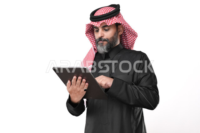 استخدام الأجهزة الحديثة في انجاز مهام الأعمال عن بعد، أداء العمل بدقة واتقان، تصفح مواقع التواصل الاجتماعي عن طريق الانترنت، بورتريه لرجل عربي خليجي سعودي يرتدي الزي التقليدي والشماغ يحمل بيده حهاز التابليت بايماءات التركيز والاندماج, خلفية بيضاء