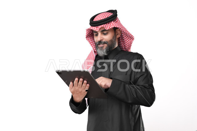 استخدام الأجهزة الحديثة في انجاز مهام الأعمال عن بعد، أداء العمل بدقة واتقان، تصفح مواقع التواصل الاجتماعي عن طريق الانترنت، بورتريه لرجل عربي خليجي سعودي يرتدي الزي التقليدي والشماغ يحمل بيده حهاز التابليت بايماءات التركيز والاندماج, خلفية بيضاء