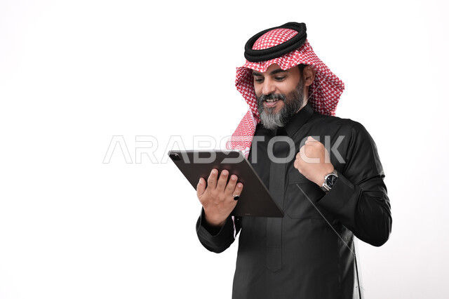 Gestures of joy for success and victory, strength and achieving goals, using modern devices to accomplish work tasks remotely, performing work with precision and mastery, portrait of an elderly Gulf Arab Saudi man wearing traditional dress and shemagh holding a tablet in his hand raising his fist up, white background