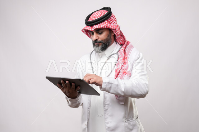 تصفح مواقع التواصل الاجتماعي عن طريق الانترنت، استخدام الأجهزة الحديثة في انجاز مهام الأعمال عن بعد، أداء العمل بدقة واتقان، بورتريه لرجل مسن عربي خليجي سعودي يرتدي الزي التقليدي والشماغ يحمل بيده حهاز التابليت بايماءات التركيز والاندماج, خلفية بيضاء
