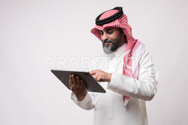 تصفح مواقع التواصل الاجتماعي عن طريق الانترنت، استخدام الأجهزة الحديثة في انجاز مهام الأعمال عن بعد، أداء العمل بدقة واتقان، بورتريه لرجل مسن عربي خليجي سعودي يرتدي الزي التقليدي والشماغ يحمل بيده حهاز التابليت بايماءات التركيز والاندماج, خلفية بيضاء