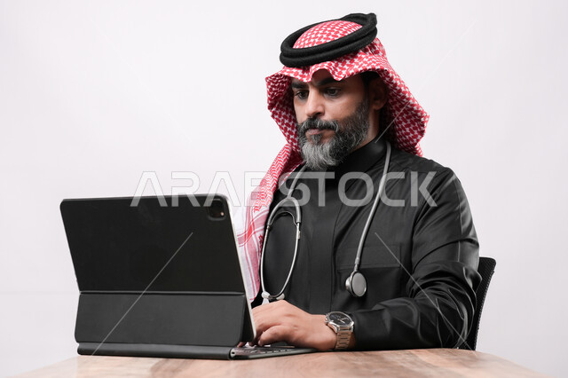 Gestures of focus and integration, browsing social networking sites via the Internet, using modern advanced devices, performing work accurately and skillfully, close-up portrait of an elderly Gulf Arab Saudi man wearing traditional dress and shemagh sitting on a chair with a tablet in front of him, concept of medicine and health care, white background