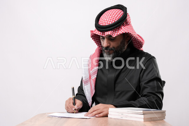 ايماءات التركيز و التمعن, مهن ووظائف مكتبية, انجاز المهام وتحقيق الاهداف, بورتريه مقرب لرجل كبير بالسن عربي خليجي سعودي يرتدي الشماغ والثوب التقليدي يجلس على الكرسي يقوم بتدوين الملاحظات على اوراق، اعداد التقارير و الملفات الخاصة بالعمل، خلفية بيضاء