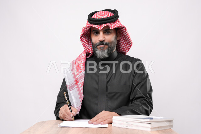 الاستعداد لكتابة و تدوين شيء ما, بيئة عمل مريحة في الشركات السعودية، بورتريه مقرب لرجل كبير بالسن عربي خليجي سعودي يرتدي الشماغ والثوب التقليدي يجلس على الكرسي  و يمسك بيده القلم بايماءات التركيز, مهن ووظائف مكتبية في المملكة العربية السعودية, خلفية بيضاء