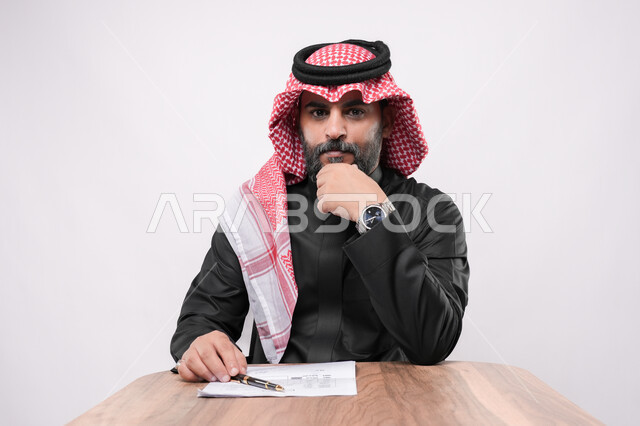 مهن ووظائف مكتبية في المملكة, الاستعداد لكتابة و تدوين شيء ما, بيئة عمل مريحة في الشركات السعودية، بورتريه مقرب لرجل كبير بالسن عربي خليجي سعودي يرتدي الشماغ والثوب التقليدي يجلس على الكرسي  و يمسك بيده القلم ينظر الى الكاميرا بايماءات التركيز, خلفية بيضاء