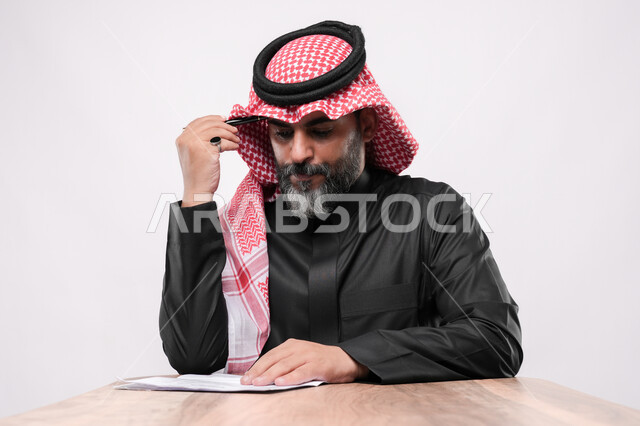 الاستعداد لكتابة و تدوين شيء ما, بيئة عمل مريحة في الشركات السعودية، بورتريه مقرب لرجل كبير بالسن عربي خليجي سعودي يرتدي الشماغ والثوب التقليدي يجلس على الكرسي  و يمسك بيده القلم بايماءات التركيز, مهن ووظائف مكتبية في المملكة العربية السعودية, خلفية بيضاء