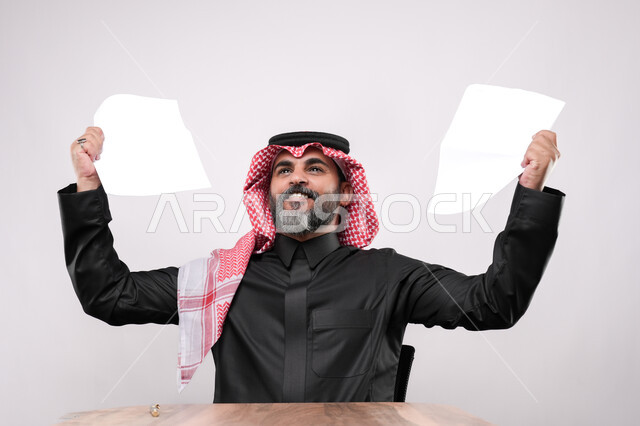 التقارير الخاصة بالعمل، بورتريه مقرب لرجل كبير بالسن عربي خليجي سعودي يرتدي الثوب التقليدي والشماغ ينظر الى الاعلى ويرفع بيده مجموعة من الاوراق البيضاء, ايماءات السعادة والفرح لتحقيق الاهداف، خلفية بيضاء