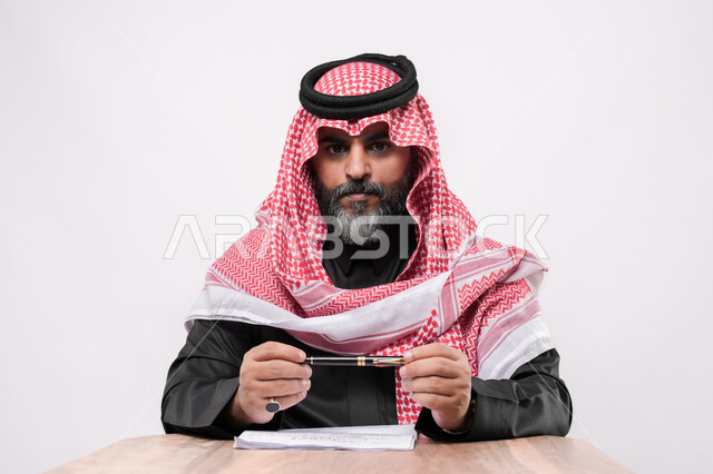 مهن ووظائف مكتبية في المملكة, الاستعداد لكتابة و تدوين شيء ما, بيئة عمل مريحة في الشركات السعودية، بورتريه مقرب لرجل كبير بالسن عربي خليجي سعودي يرتدي الشماغ والثوب التقليدي يجلس على الكرسي  و يمسك بيده القلم ينظر الى الكاميرا بايماءات التركيز, خلفية بيضاء
