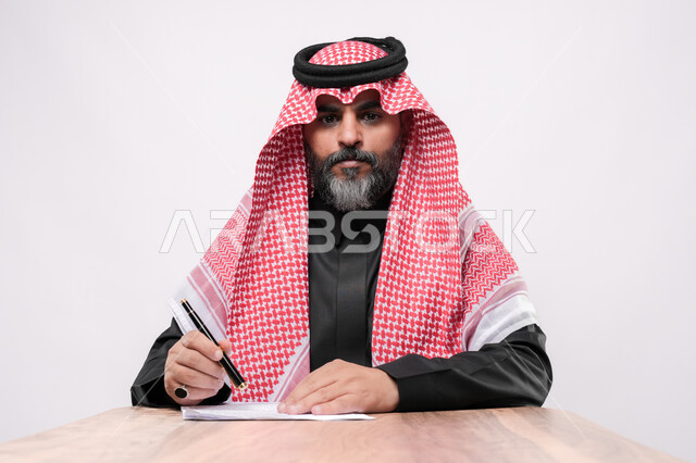 بيئة عمل مريحة في الشركات السعودية، الاستعداد لكتابة و تدوين شيء ما, بورتريه مقرب لرجل كبير بالسن عربي خليجي سعودي يرتدي الشماغ والثوب التقليدي يجلس على الكرسي  و يمسك بيده القلم بايماءات التركيز, مهن ووظائف مكتبية في المملكة العربية السعودية, خلفية بيضاء