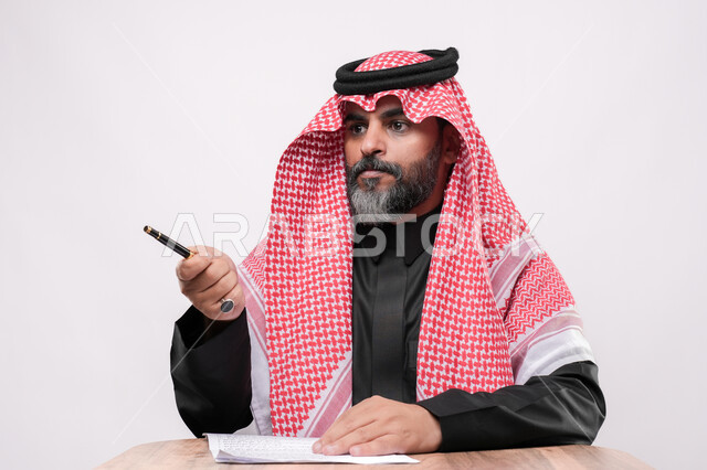 بيئة عمل مريحة في الشركات السعودية، الاستعداد لكتابة و تدوين شيء ما, بورتريه مقرب لرجل كبير بالسن عربي خليجي سعودي يرتدي الشماغ والثوب التقليدي يجلس على الكرسي  و يمسك بيده القلم بايماءات التركيز, مهن ووظائف مكتبية في المملكة العربية السعودية, خلفية بيضاء