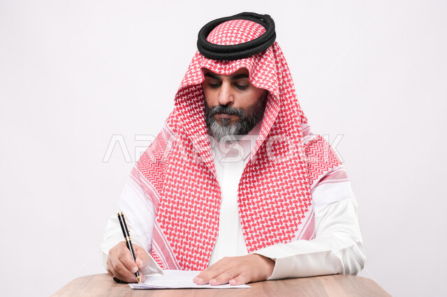 ايماءات التركيز و التمعن, مهن ووظائف مكتبية, انجاز المهام وتحقيق الاهداف, بورتريه مقرب لرجل كبير بالسن عربي خليجي سعودي يرتدي الشماغ والثوب التقليدي يجلس على الكرسي يمسك بيده قلم يقوم بتدوين الملاحظات على اوراق، اعداد التقارير و الملفات الخاصة بالعمل، خلفية بيضاء