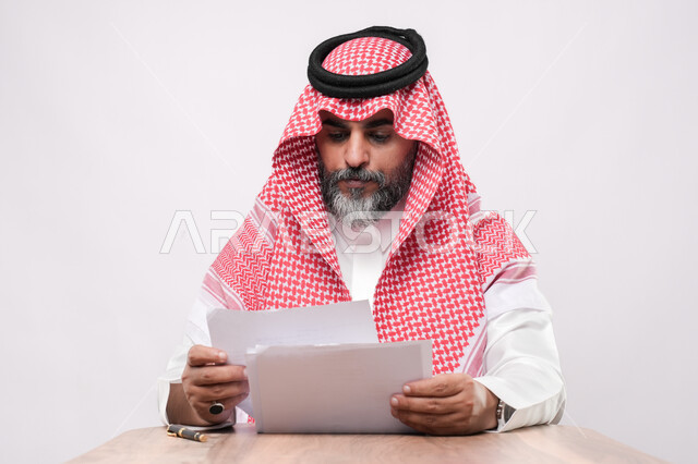 متابعة ومراجعة الخطط والمشاريع المكتبية، اعداد التقارير الخاصة بالعمل، بورتريه مقرب لرجل كبير بالسن عربي خليجي سعودي يرتدي الثوب التقليدي والشماغ ينظر الى الاوراق البيضاء, التدقيق في قراءة الملاحظات، خلفية بيضاء