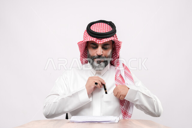 مراجعة ومتابعة الخطط والمشاريع المكتبية، اعداد التقارير الخاصة بالعمل، بورتريه مقرب لرجل كبير بالسن عربي خليجي سعودي يرتدي الثوب التقليدي والشماغ يقوم بالتدقيق في قراءة الملاحظات، خلفية بيضاء