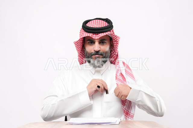 مراجعة ومتابعة الخطط والمشاريع المكتبية، اعداد التقارير الخاصة بالعمل، بورتريه مقرب لرجل كبير بالسن عربي خليجي سعودي يرتدي الثوب التقليدي والشماغ يقوم بالتدقيق في قراءة الملاحظات، خلفية بيضاء
