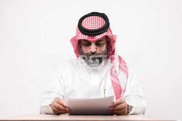 اعداد التقارير الخاصة بالعمل، بورتريه مقرب لرجل كبير بالسن عربي خليجي سعودي يرتدي الثوب التقليدي والشماغ يحمل بيده مجموعة من الاوراق يقوم بقراءة الملاحظات, مراجعة ومتابعة الخطط والمشاريع المكتبة, ايماءات التمعن والتركيز، خلفية بيضاء