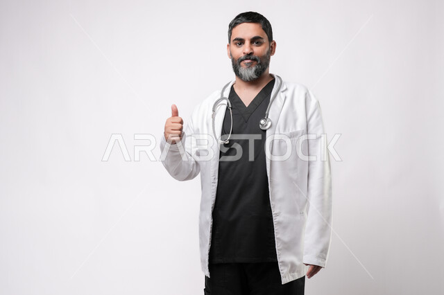 مفهوم الامتياز والجودة, الاستعداد لتقديم الخدمات للمرضى بكل كفاءه، مفهوم الطب والرعاية الصحية، بورتريه لطبيب كبير بالسن عربي خليجي يرتدي المعطف الطبي والسماعه يقوم برفع اصبع الابهام للاعلى بتعابير القبول والموافقة، مهن ووظائف طبية في القطاع الصحي, خلفية بيضاء