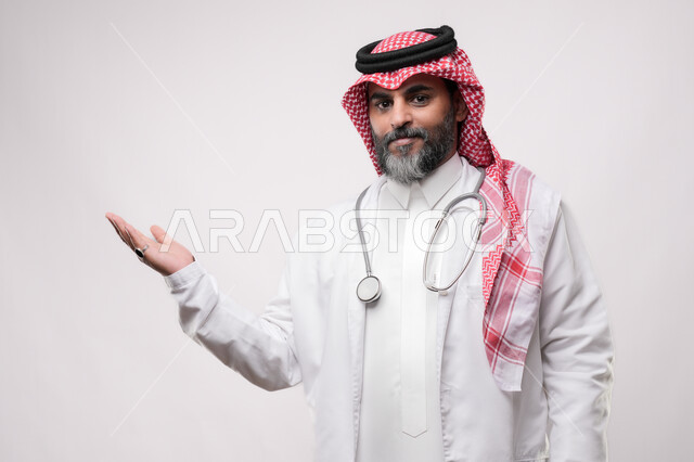 الترحيب والاستقبال ببشاشة، بورتريه لطبيب مسن عربي سعودي خليجي يرتدي الشماغ والثوب التقليدي والسماعه ينظر الى الكاميرا يفتح كفه بتعابير رؤية شخص ما، الاستعداد لتقديم الخدمات للمرضى بكل كفاءه، مفهوم الطب والرعاية الصحية، الإشارة نحو هدف معين، خلفية بيضاء